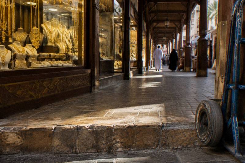 Low and close on the worn stone threshold of the Gold Souk in Deira morning light slanting into a covered arcade of jewellery windows, polished brass glinting, a scuffed delivery trolley wheel in frame.