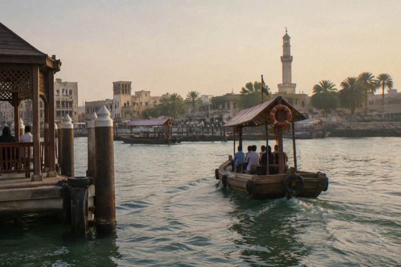 An abra pulling away from the Bur Dubai bank across the green water of Dubai Creek toward Deira in hazy afternoon light.